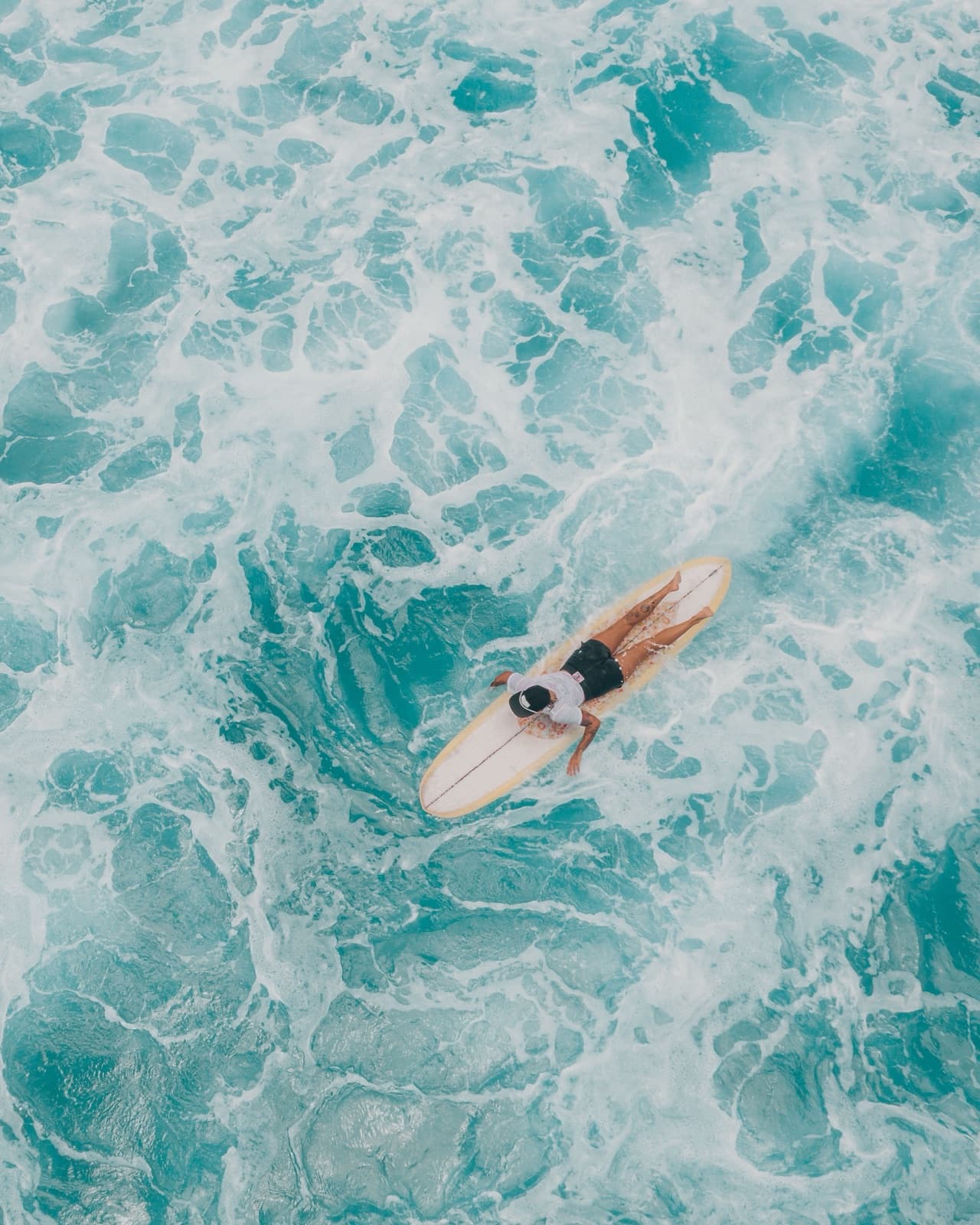 A surfer paddling through turquoise water, seen from above