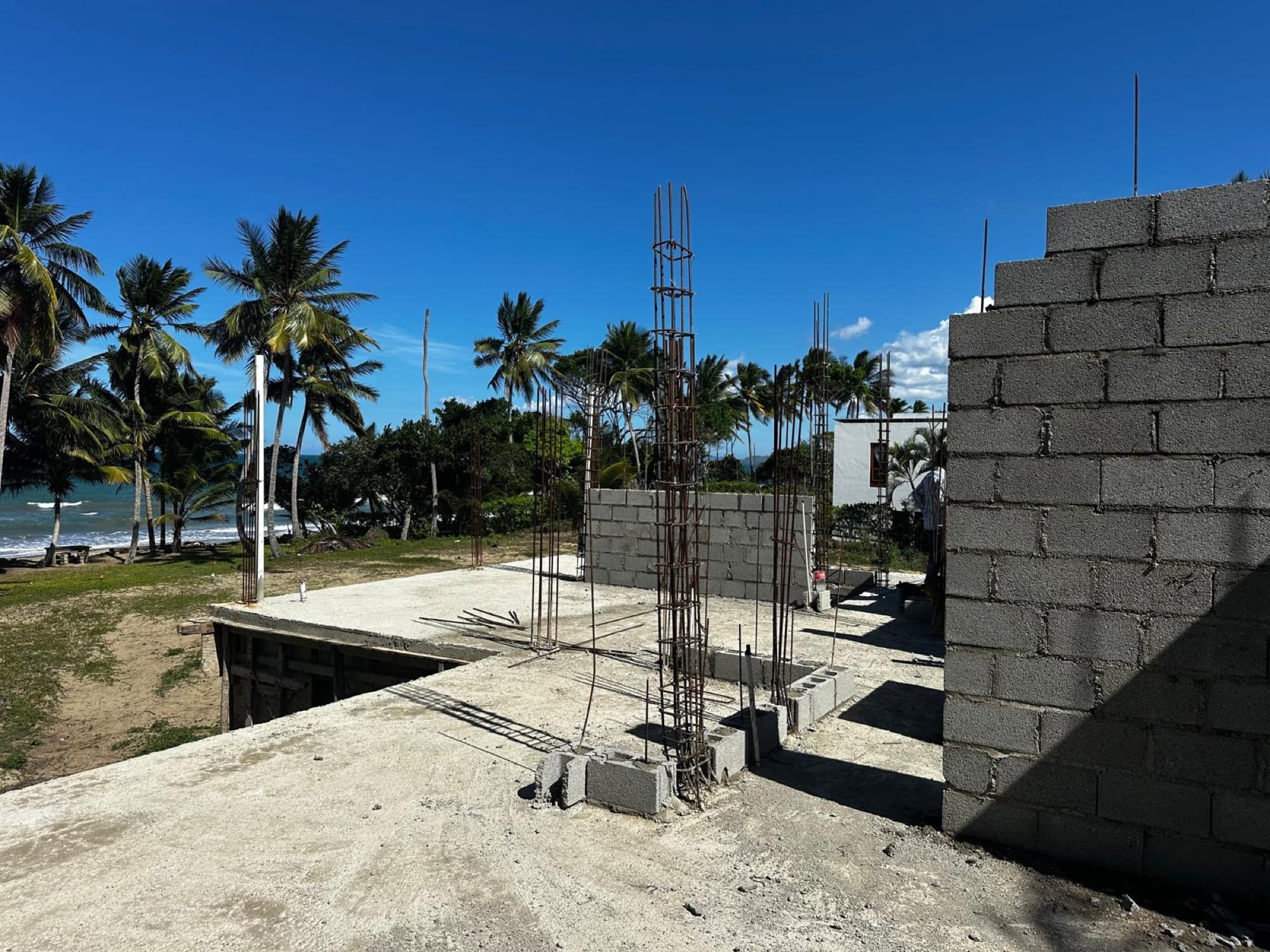 On the upper slab: column reinforcement standing and the first upper-level block walls laid, the sea in view.